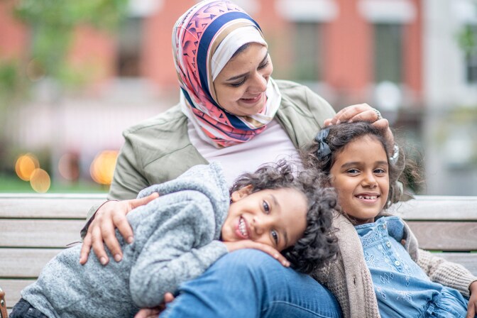 A smiling woman wearing a hijab sits with two smiling children on a bench. | stock