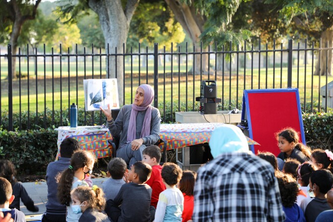 A woman wearing a headscarf holds up a picture book and reads to a group of children sat down in front of her. The reading is taking place in an outdoor area. Behind the woman and group of children is a black iron fence. Beyond that is an open area of green grass and tall trees.