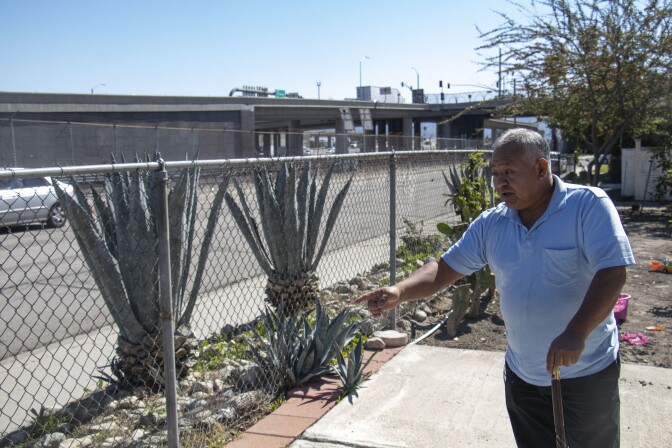 Benjamin Luna stands holding a cane and pointing toward a fence.