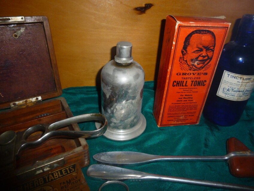Vintage medical artifacts are displayed on a dark green satin cloth. Reflex hammers, tinctures and medicine boxes are arranged together. 