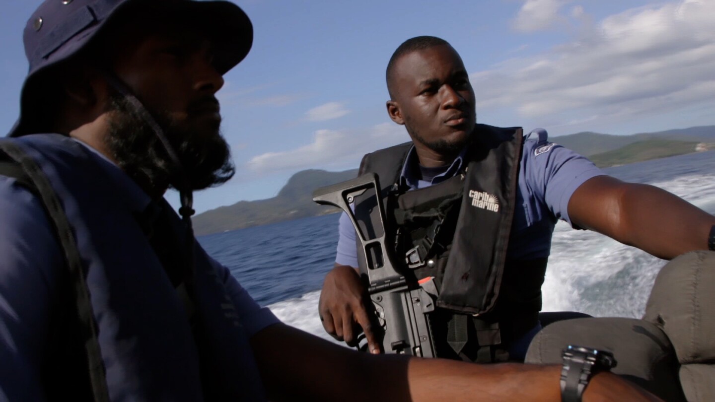 Two men stand on a boat, with one of them handling a rifle. | From "Foreign Correspondent: Pirates of the Caribbean"
