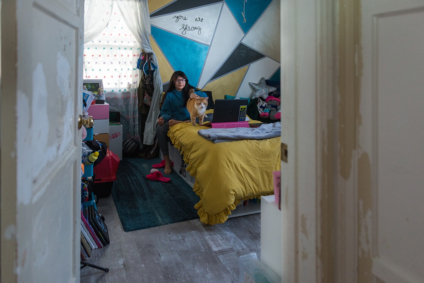Rhianna plays with her cat in her bedroom. She's sitting on a mustard yellow blanket with a laptop on her bed. Behind her is a geometric painted wall with triangles painted in various blues and yellow.
