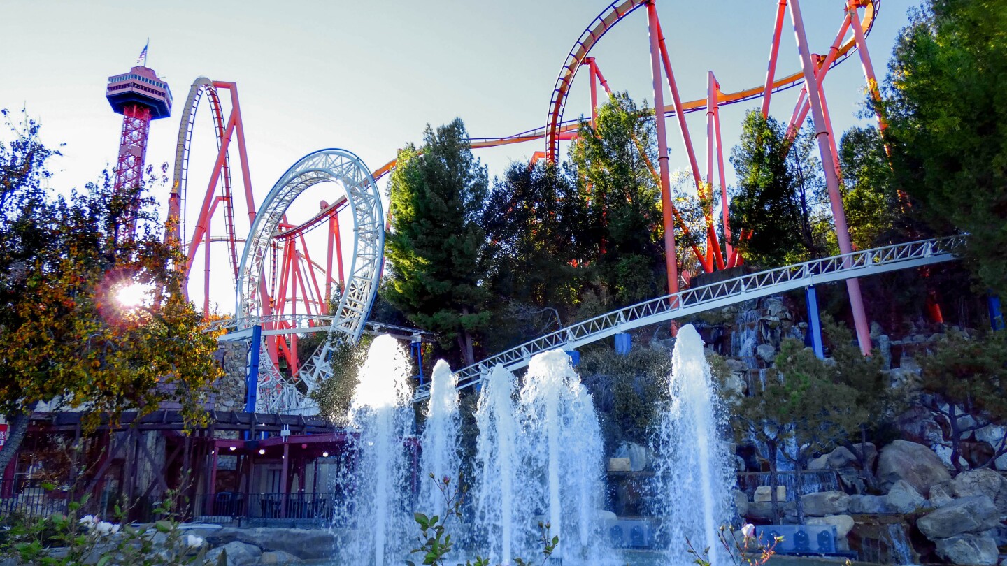 The Revolution Classic rollercoaster tracks are white and loop at a certain point. Behind is another rollercoaster with red tracks and is taller than the Revolution Classic. A red observation tower can be seen in the near distance. In the foreground, water spurts upwards in a fountain/pond.
