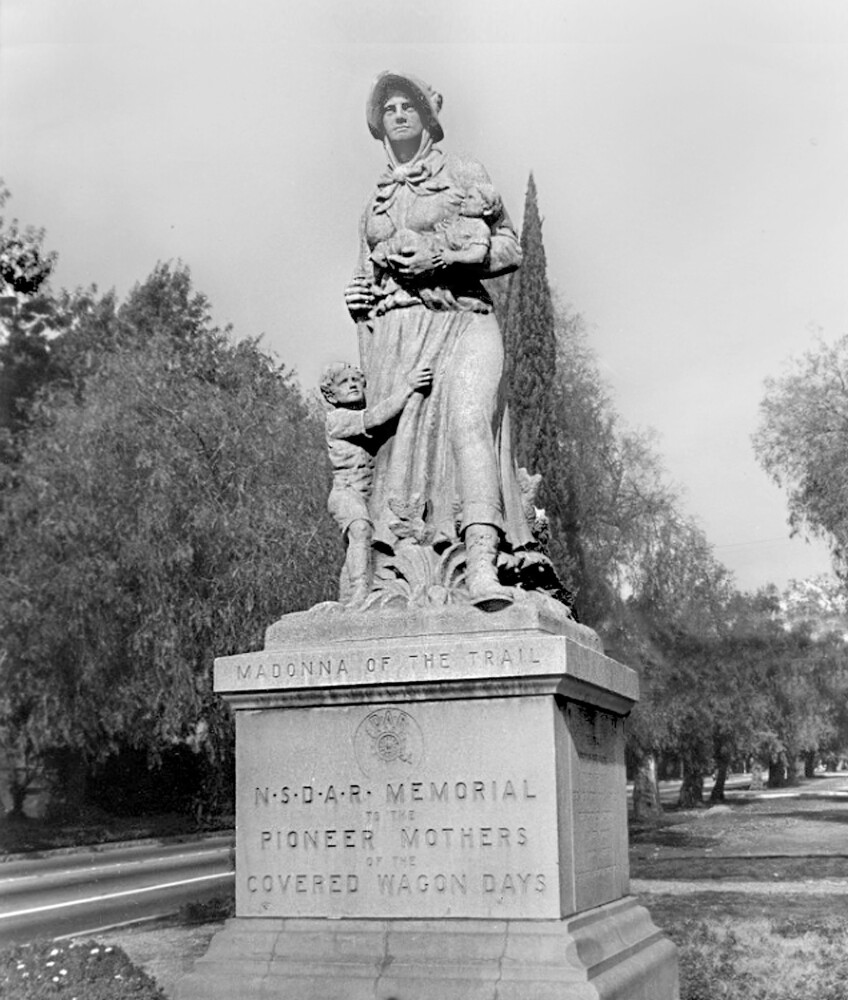 A black and white photo of a stone sculpture of a woman, dressed in a attire that old Californian pioneers would wear. She looks sternly off into the distance while cradling a baby in one arm and a rifle in the other. A young boy clutches on her leg, tugging at the "fabric" of her dress. The sculpture stands on a base that has engraved: "Madonna of the Trail," and below that, "NDSAR Memorial to the pioneer mothers of the covered wagon days."