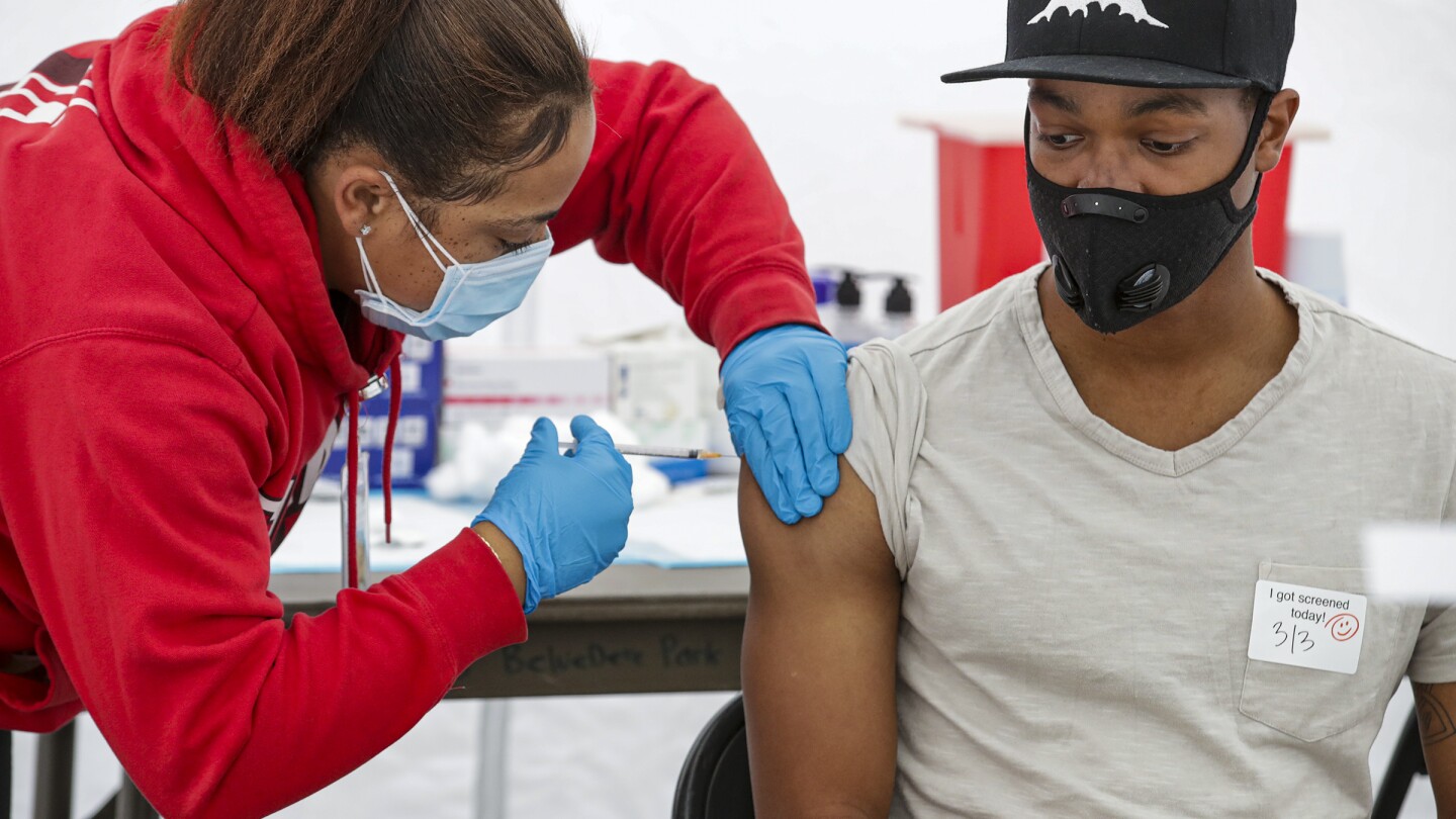 A person in a red sweatshirt administers COVID-19 vaccine to a young Black man. Both are wearing face masks.