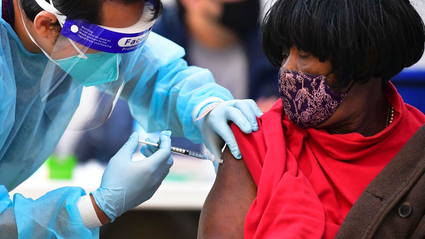 A nurse in blue personal protective gear administers the Moderna Covid-19 vaccine to a woman in a red shirt.