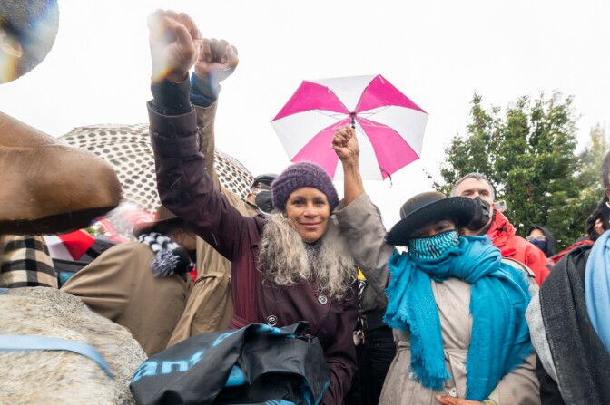 Dana King, who is wearing a purple knit beanie over her curly gray hair, stands next to Congresswoman Barbara Lee, who is wearing a bright blue scarf around her neck and a black brimmed hat. Both of the women have their fists raised in the air. Attendees around them are holding umbrellas. They sky above is overcast and gray