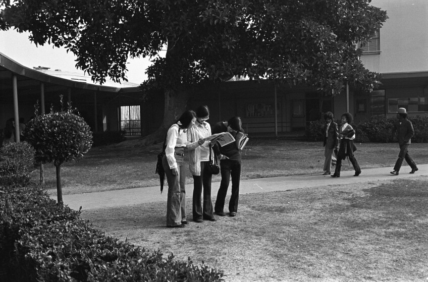 A black and white photo of a group of three Asian American students gathered on a lawn at Susan Miller Senior Dorsey High. circa 1972. They're all gathered around the student in the middle, who is holding open a magazine or newspaper of some sort. Beyond, a group of Black students walk on the sidewalk with books in hand. 