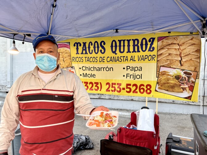 Raul Quiroz is wearing a striped red and cream collared shirt and a royal blue cap over his head. He's also wearing a surgical face mask. Behind him is a yellow sign that reads, "Tacos Quiroz: Ricos tacos de canasta al vapor." Below are the following presented as a bulleted list: "chicharr´on, papa, mole and frijol." Below that is a phone number: (323) 253-5268. Raul is holding a styrofoam plate containing tacos.