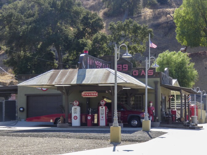 A 1950s-era rustic roadside gas station stands among a dirt patch. The roofline reads, "Hwy 99 Gas." Below are two vintage gas pumps and a red truck parked nearby.