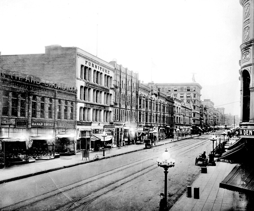 Black and white archival photo of a Los Angeles city street in the early 1900s. Ornate street lamps line the street and tracks run down a street. 