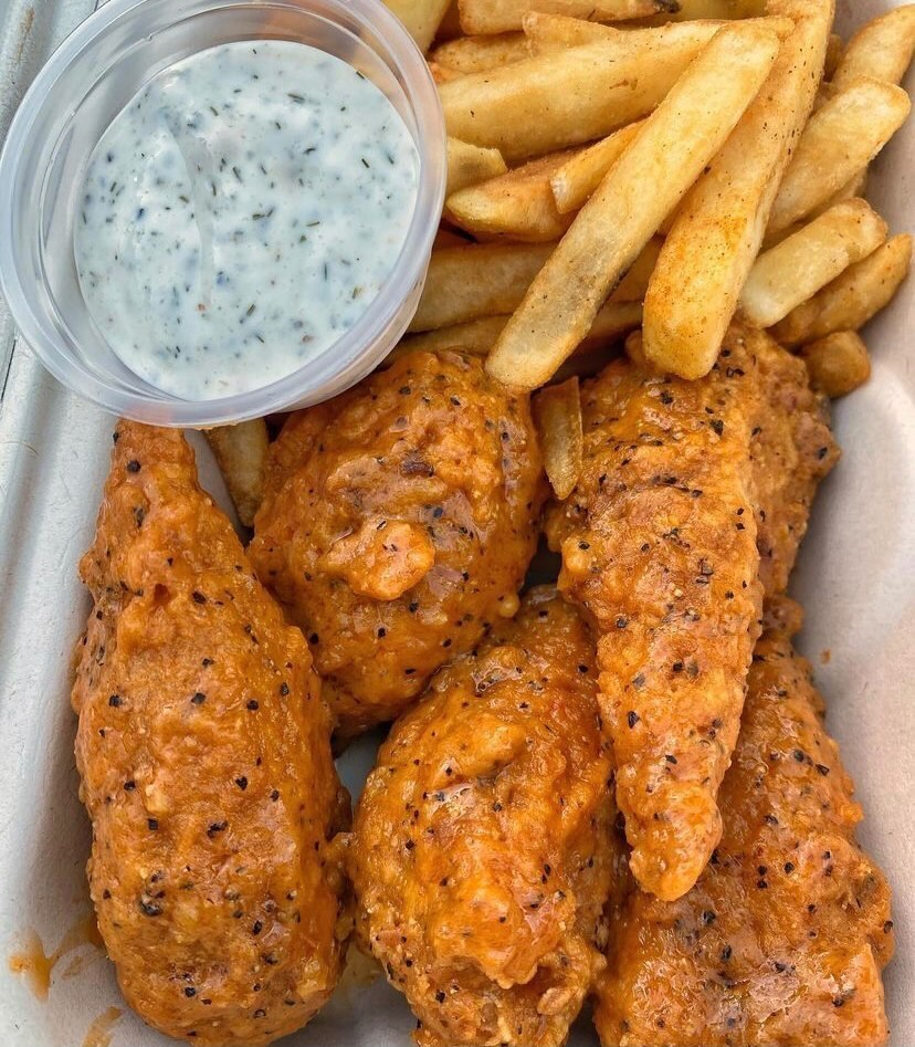 Fried and crispy vegan chicken seasoned with black pepper and smothered in an orange sauce sit in a paper takeout box with a side of fries and a white dipping sauce.