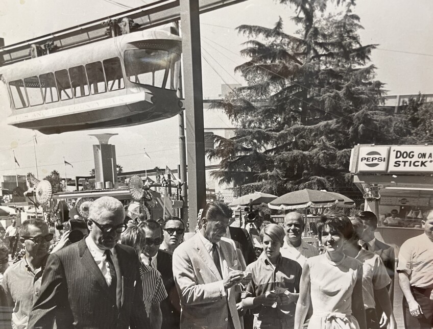 A black and white photo of the Los Angeles County Fair. A monorail travels above the crowds. Below, former California governor Ronald Reagan and his wife Nancy Reagan walk amongst the crowd. Ronald Reagan is seen signing a piece of paper while a young boy waits next to him.
