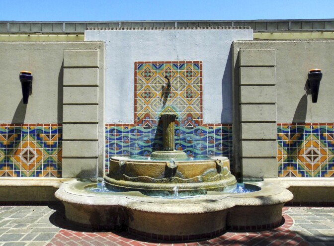 An outdoor water fountain spews out water that trickles down its two tiers. The fountain is attached to a wall decorated in brightly colored tiles, forming geometric shapes and patterns.