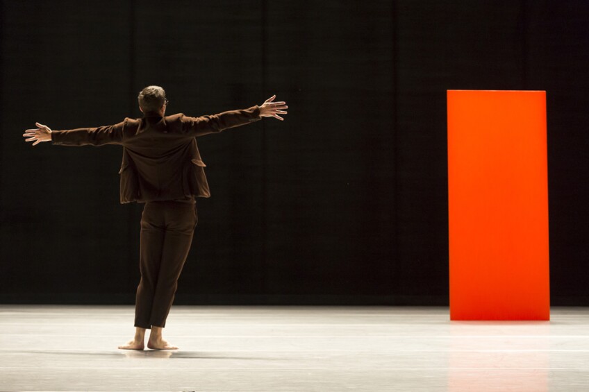 Image of the back of a bare foot man in suit standing on stage with arms outstretched.