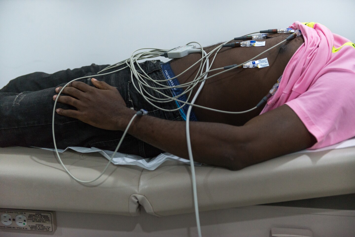 A man lays on a hospital bench with his pink shirt pulled up, exposing his torso. On his torso are various wires attached to his chest.