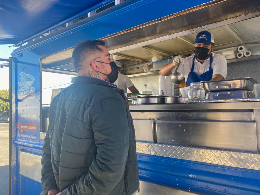 Chef Jos´e Figueroa stands in front of a food truck, watching his order as its being prepared by an employee inside the truck. The employee is pouring a brown broth into a styrofoam cup with a large metal ladle. The food truck is blue and a small logo that reads, "Los Chilos" over an illustration of a shark is posted on the left side of the food truck window.
