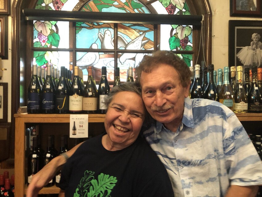 Dora Herrera and Tony Fanara pose for a picture inside Palermo. Behind them is a shelf displaying various wines. Behind that is a stain glass window depicting doves drinking and bathing in a water fountain.  