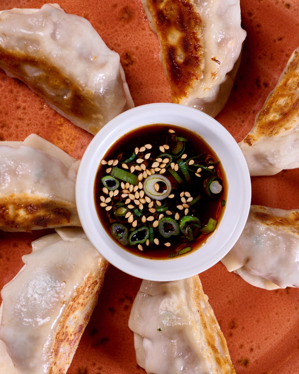 A flat lay photo of a plate of pan fried dumplings arranged in a circle around a small holder for dipping sauce. The dipping sauce is stopped with sliced green onions and sesame seeds floating on the dark liquid's surface. The dumplings and sauce are on an orange ceramic plate sitting on a dark wood surface.