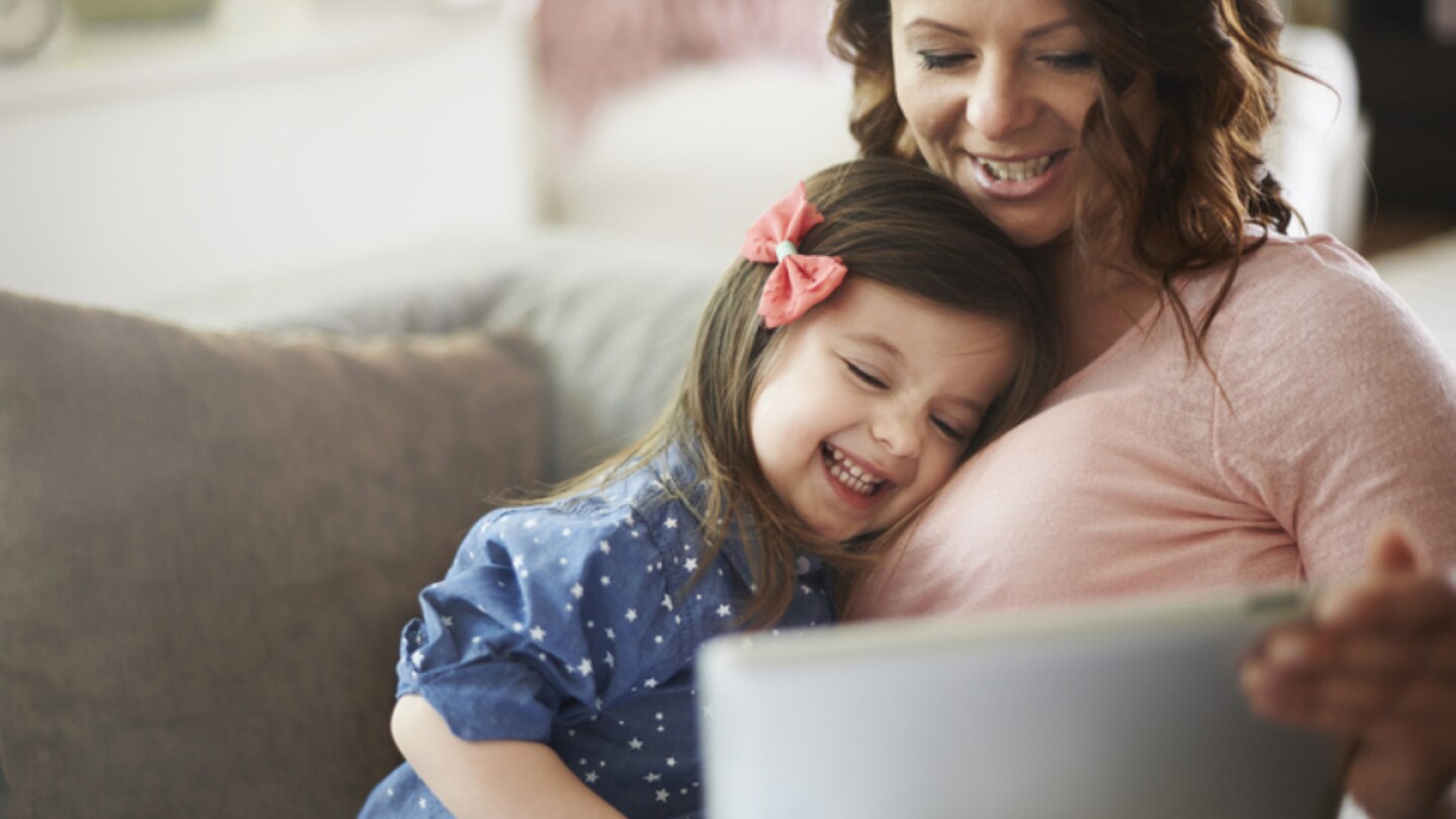 A woman and a little girl sit on a couch in front of a laptop.