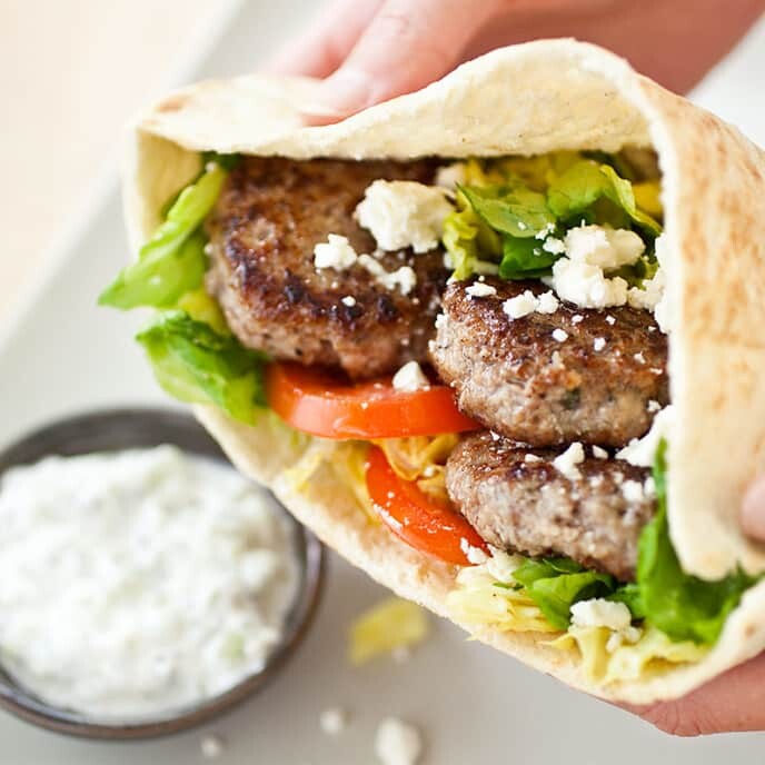 A pita sandwich filled with tomatoes, small clumps of feta cheese, lettuce and small patties of meat, with a white sauce in the background.