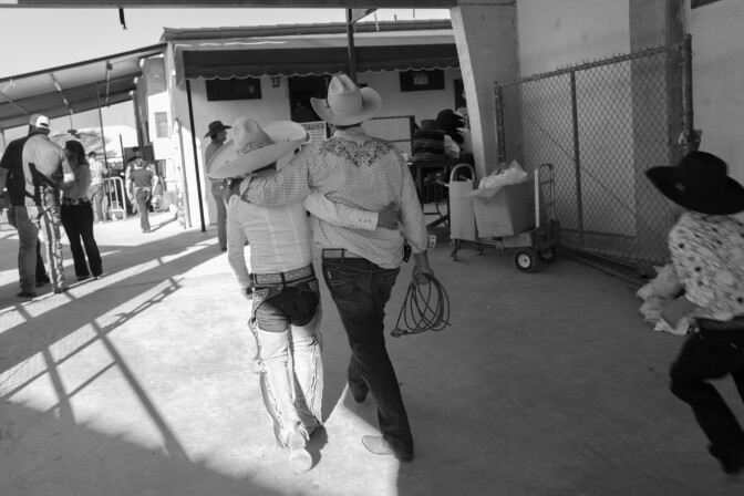 Dos hombres con camisas bordadas, chaparreras de cuero y pantalones a juego caminan de la mano. 