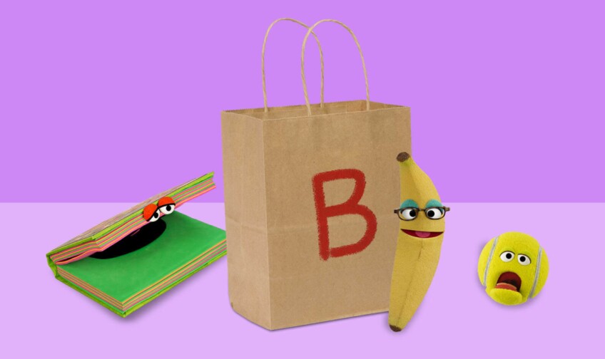Un cuaderno, plátano y pelota con ojos de caricatura están tendidos al lado de una bolsa de papel con la letra "B"