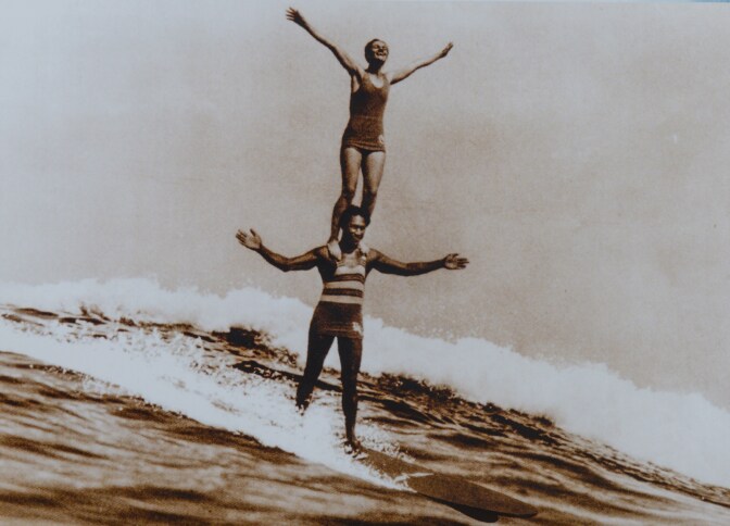 A sepia-toned photo of Duke Kahanamoku surfing a wave while Viola Hartman stands on his shoulders. The two have their arms outstretched as they ride the wave.