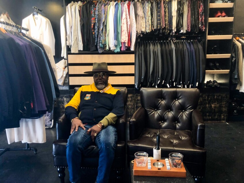 Khnum Alexander of Swank Men's Fashion sits on a black cushioned arm chair. All around him are racks of mens wear from blazers and coats to dress shirts and trousers. 