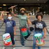 Three young Black Lives Matter protestors hold their drumsticks high in a fist with a drum tied around their waist.