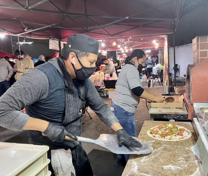 A man wearing an apron prepares pizzas at an outdoor market. He's holding a metal pizza peel over a floured surface with an uncooked pizza sitting atop.