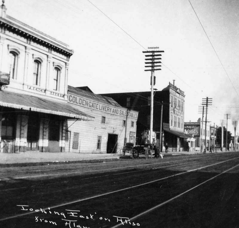 The French district in Los Angeles is shown looking east on Aliso Street from Alameda Street. The photo includes a French bakery and the Golden Gate Livery and Sale Stables are shown.