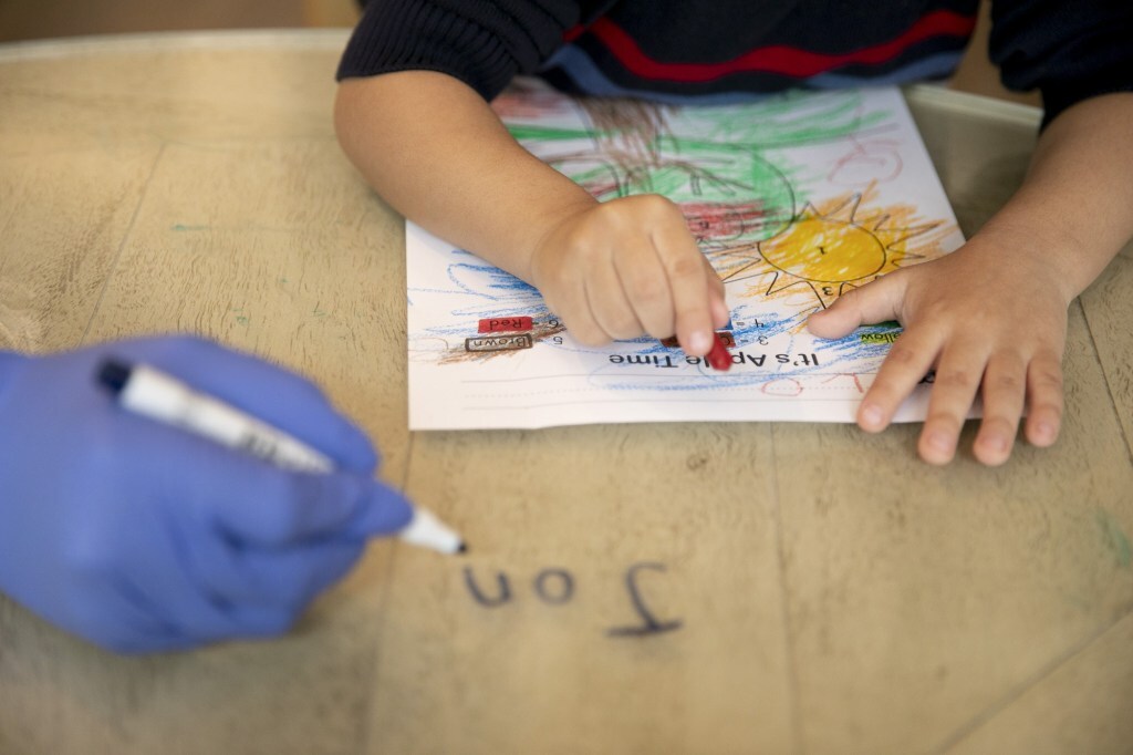 Jonathan's hands are on the table, coloring a sheet of paper. The paper has a sun on it. Off-camera, a gloved hand holding a marker writes, "Jon" on the table.