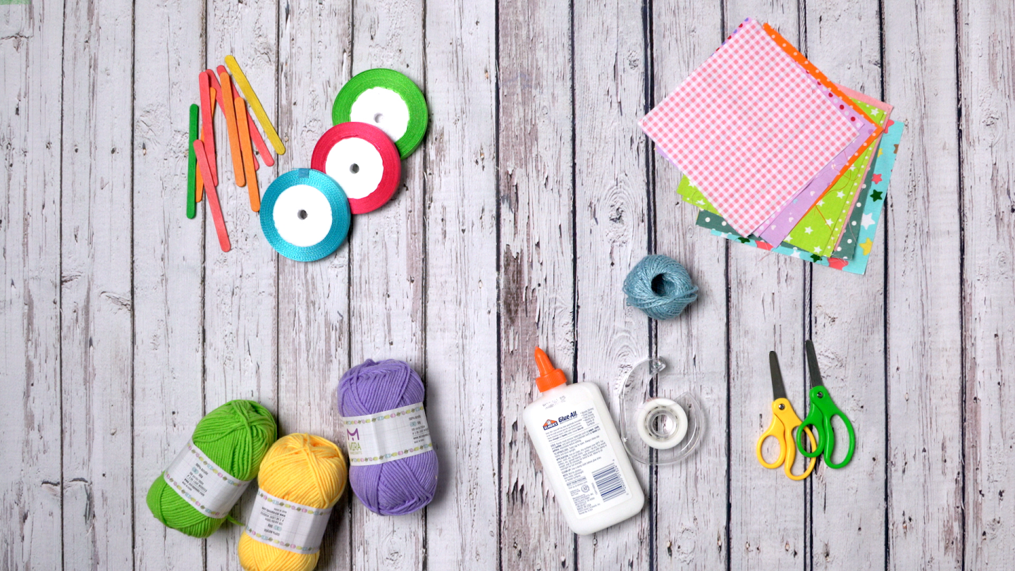 Balls of yarn, tape, scissors, a glue container, popsicle sticks and small pieces of fabric are arranged on a table.