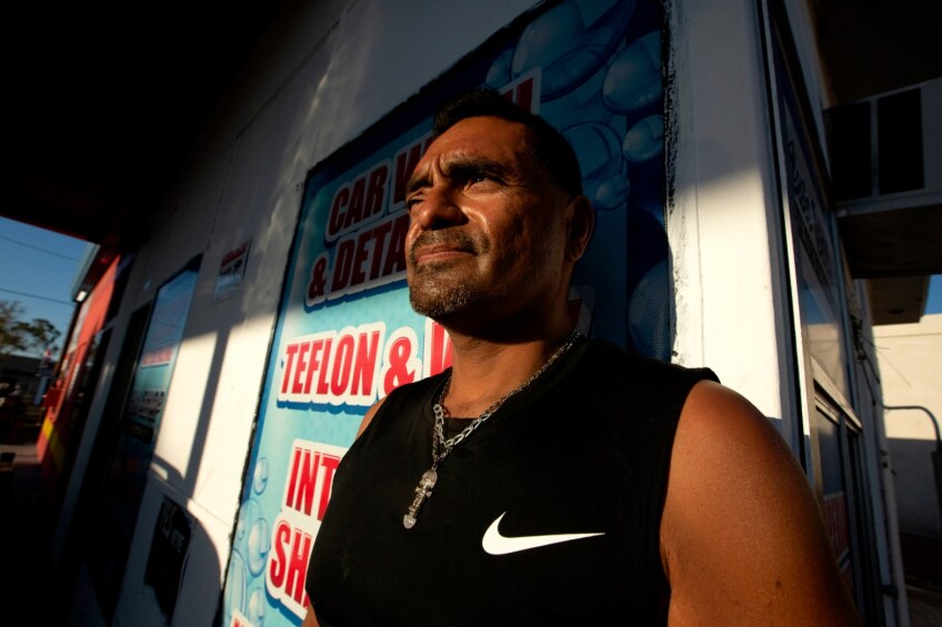 Micheal Torres sits for a portrait inside the auto shop where he works in El Centro on April 29, 2021. He has brown skin, wears a steel cross around his neck, a sleeveless black top and black, short hair and a goatee.