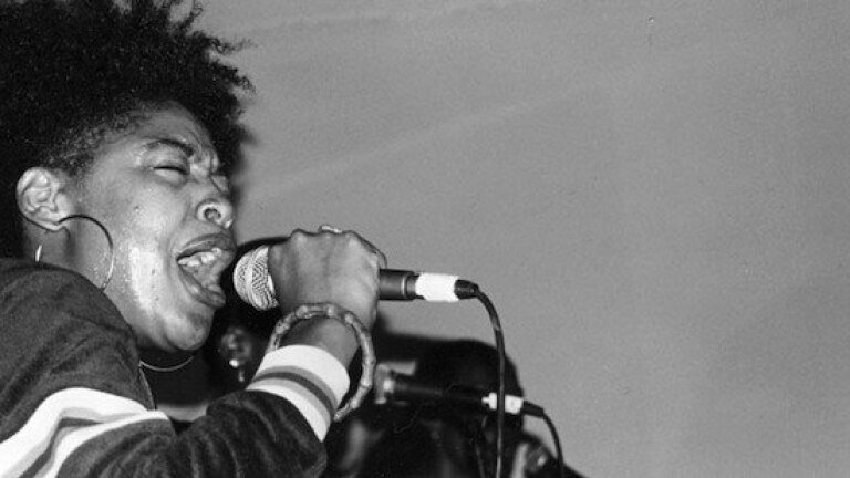 A black and white hpoto of Medusa The Gangsta Goddess, a young Black woman with her hair in an updo and wearing large hoop earrings. She is holding a microphone up to her mouth as she performs passionately into it.