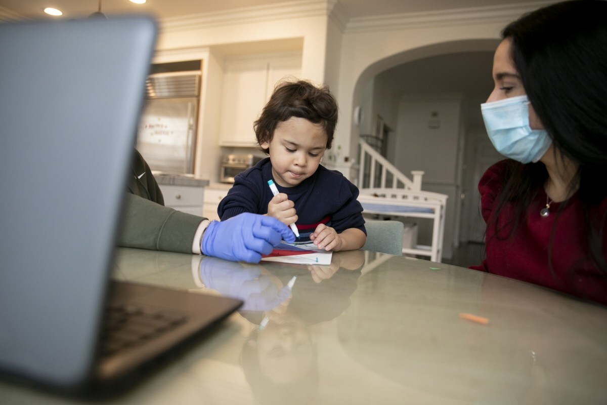 Jonathan, 4, holds a marker to draw on a paper set in front of him on a table. Next to him is his mother, watching him draw. She's wearing a face mask.
