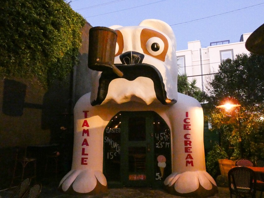 A building shaped like a giant bull dog smoking a pipe sits between a vine-covered wall and patio seating. On each of the dogs legs are words painted in red. On the left leg is the word, "tamale." On the right leg is the word, "ice cream." 