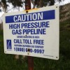 A close-up of a rusty white sign with blue text that reads "CAUTION HIGH PRESSURE GAS PIPELINE" and "BEFORE EXCAVATING IN AREA CALL TOLL FREE SOUTHERN CALIFORNIA GAS COMPANY" and a toll-free number, alongside a SoCalGas logo in blue. In the background is a chain link fence, a leafy tree, and a paved road on a gloomy day.