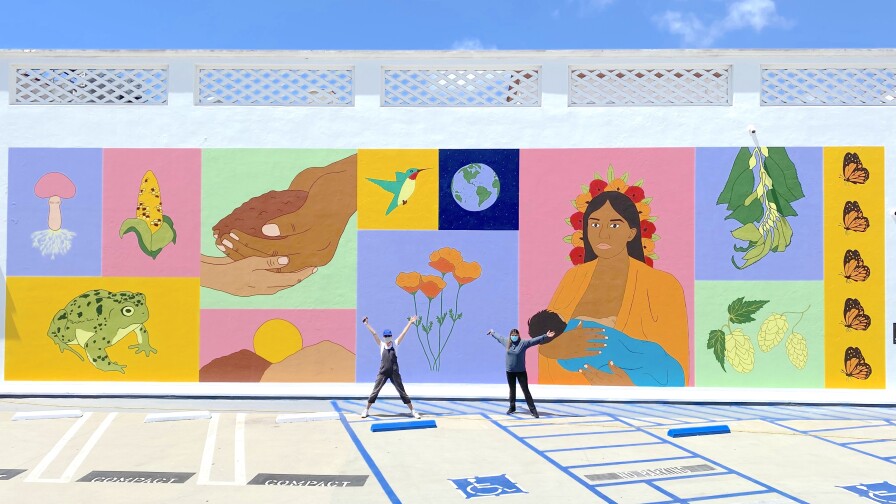 Two women stand in front of a large and colorful mural. The mural consists of images segregated in their own square, each with their own color block background. One of the squares is yellow with a frog. Another square is pink with the image of a dark-skinned woman with long hair and holding a baby. Other imagery on the tiles include California poppies, corn, a hummingbird, the earth, butterflies and two hands holding soil.