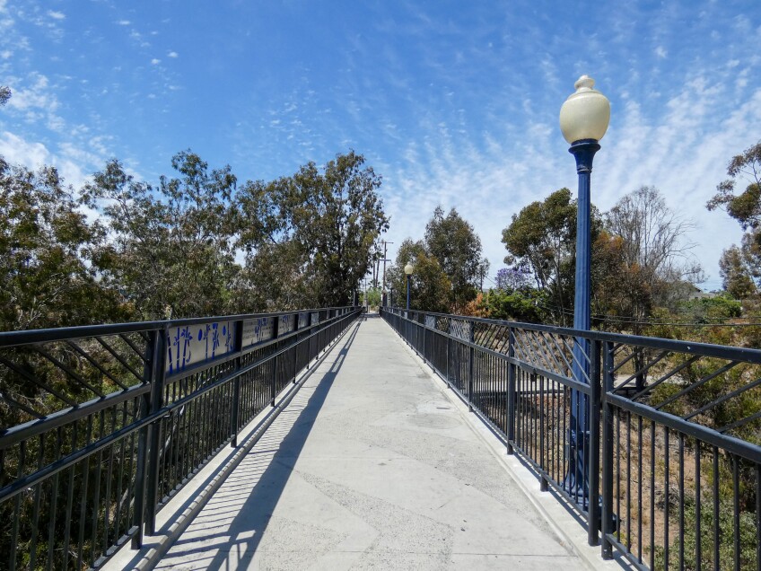 A photo of a long, concrete bridge with steel railings on either side. The bridge crosses through a green forest area. Two blue lamp posts are attached to the right side of the bridge. 