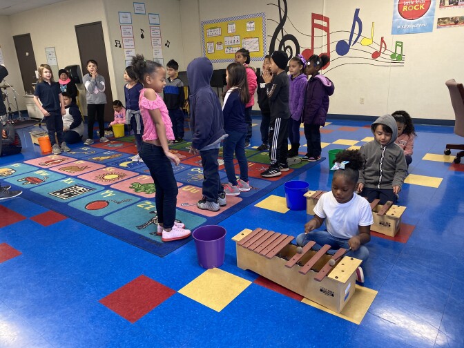 Students assemble in a classroom with some lining up while others play music on a xylophone.