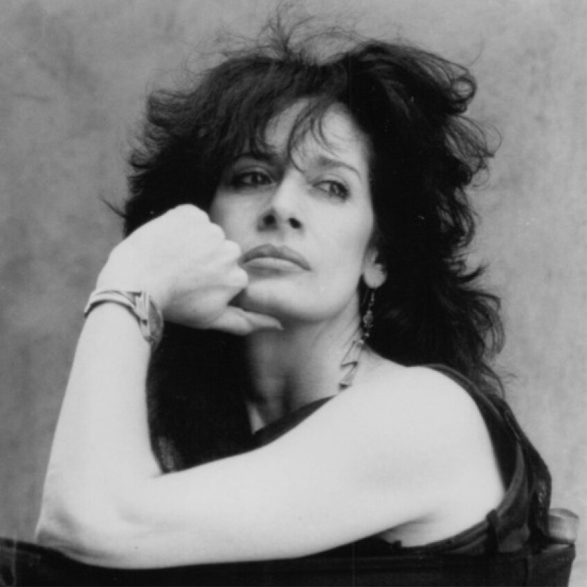 A black and white photo of Penelope Spheeris. She has long dark hair styled in a blowout. She is resting her elbow on the back of a chair and rests her chin on her hand as she looks off to the sidd. 