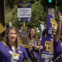 Women strikers wear purple and hold black and yellow signs that read in large block print "PATIENT CARE IS IN CRISIS" and "PATIENT NEEDS NOT CORPORATE GREED"