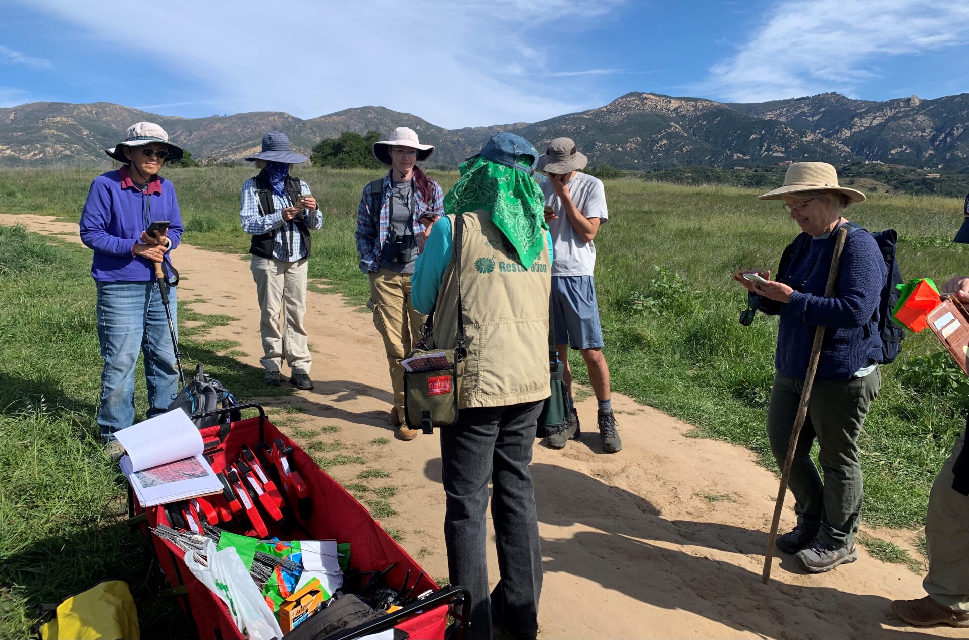 A group of volunteers gather in a circle on a dirt footpath that cuts through a green open field of grass. Beyond, mountains carve the sky. Some volunteers are taking notes on their phone, as their attention is directed towards an individual wearing a beige vest. On the back, it reads, "Channel Islands Restoration" in green. Next to them is a red wagon with various materials related to restoration.