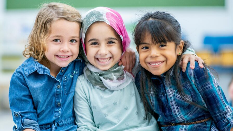 Three little girls, one blond, another wearing hijab and another with black hair, hug each other and smile.