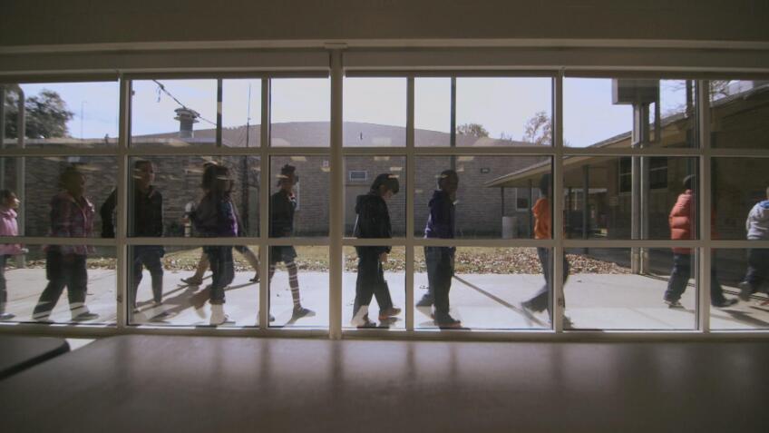 Children walk in a line outside a building with tall windows.