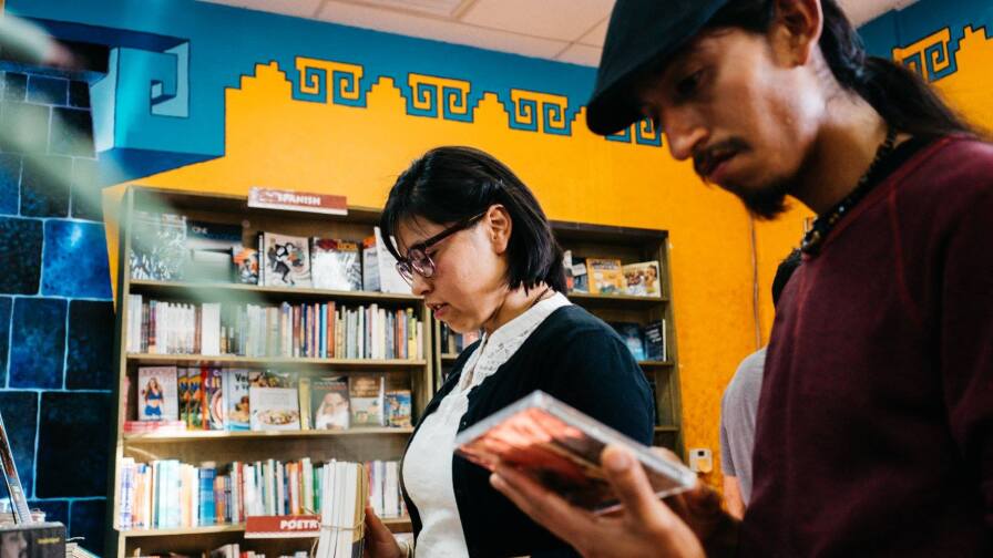 Two people standing side by side inspect a book and CD, respectively. Behind them is a bookcase pushed up against a vibrant yellow wall with teal blue detailing at the top.