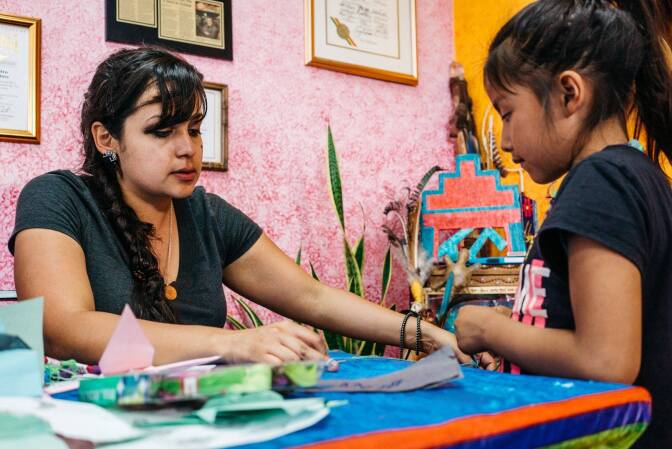 A young woman sits at a table and speaks to a child. The two are sitting in a vibrant environment with a bright pink and white wall on one side of them and a marigold yellow wall on the other.