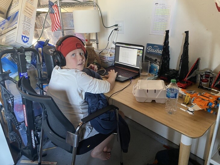 A young boy in a red hat works at a desk with a laptop.